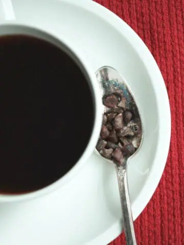 overhead image of coffee in mug on saucer next to spoon holding cacao nibs