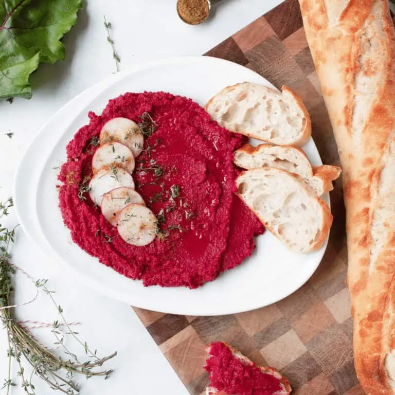 overhead image of savory roasted beet hummus on plate with baguette to one side and herbs on the other