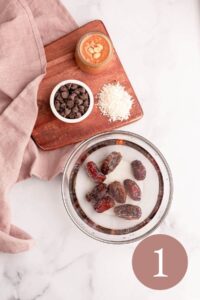 step 1: overhead image of peanut butter dates soaking on counter next to chocolate chips, shredded unsweetened coconut, and jar of peanut butter