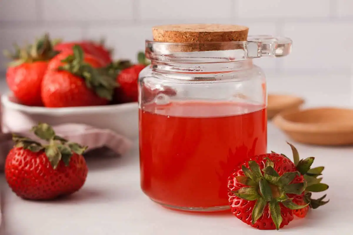 side image of strawberry syrup in glass jar with strawberry tops in the foreground and whole strawberries in the background