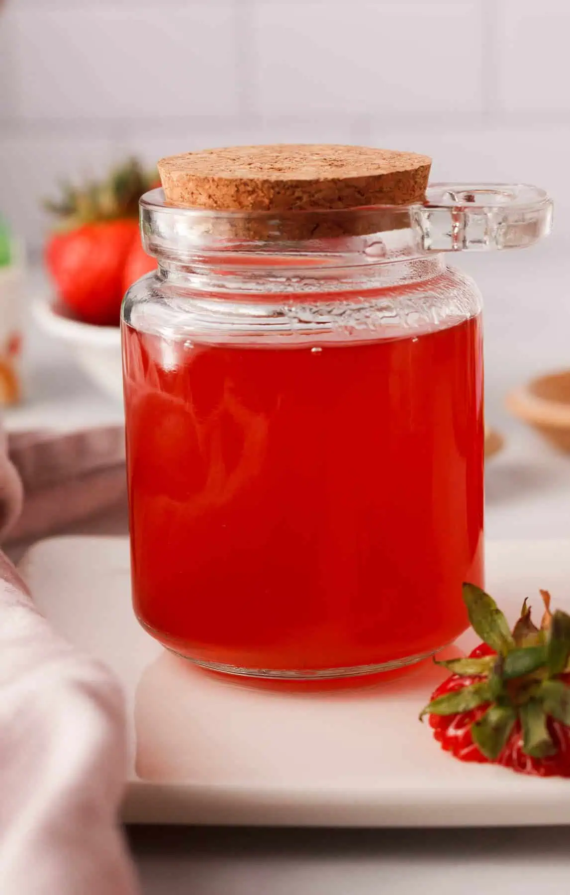 closeup of strawberry syrup in glass jar with corked lid and strawberry tops propped against the side