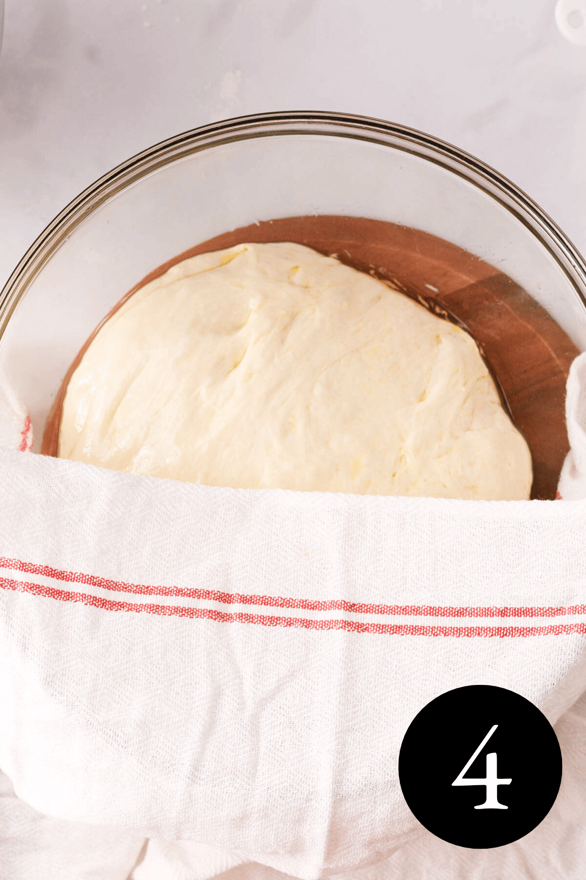 overhead image of focaccia dough in glass bowl with towel partially covering the top in preparation of the first rise