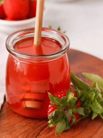 closeup of strawberry basil simple syrup in glass jar with honey dipper in jar and strawberries and basil in foreground and background
