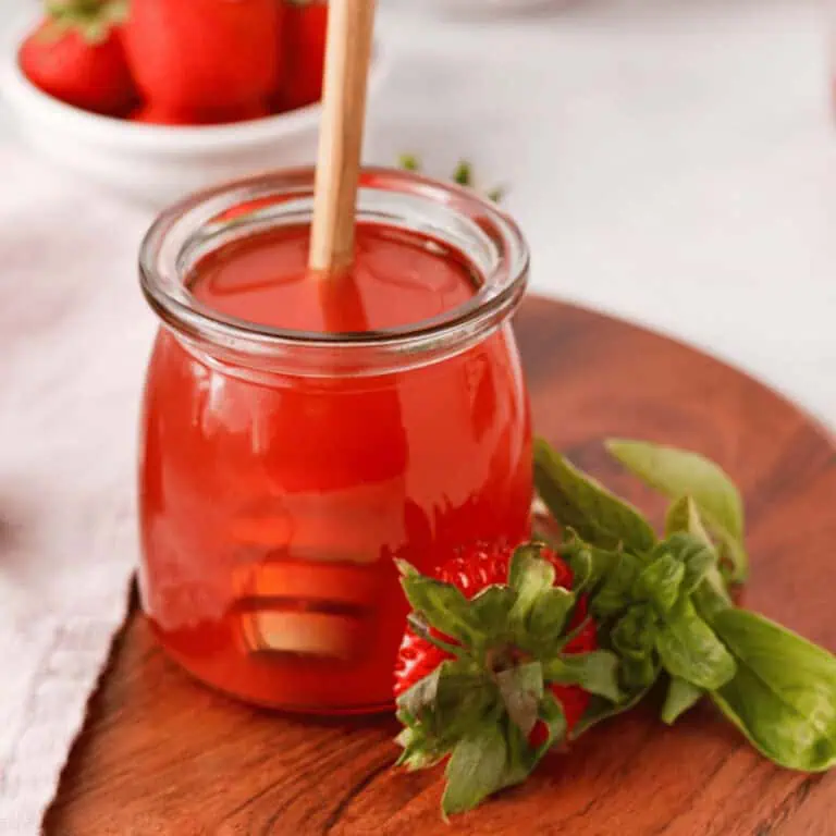closeup of strawberry basil simple syrup in glass jar with honey dipper in jar and strawberries and basil in foreground and background