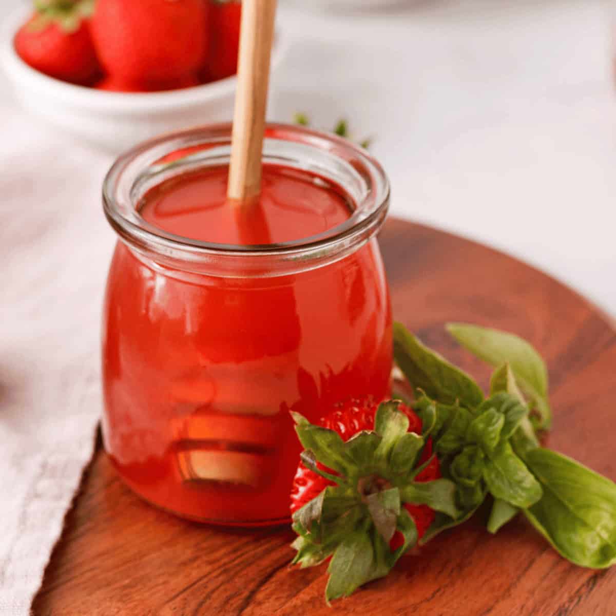closeup of strawberry basil syrup in jar with honey dipper and strawberry top and fresh basil propped against side