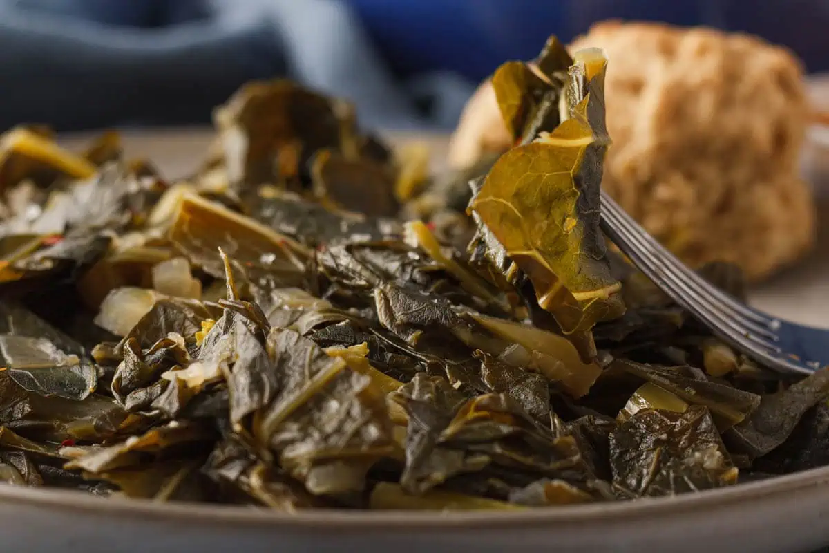 closeup of fork holding up a piece of miso collard green with plate of greens and biscuit in background