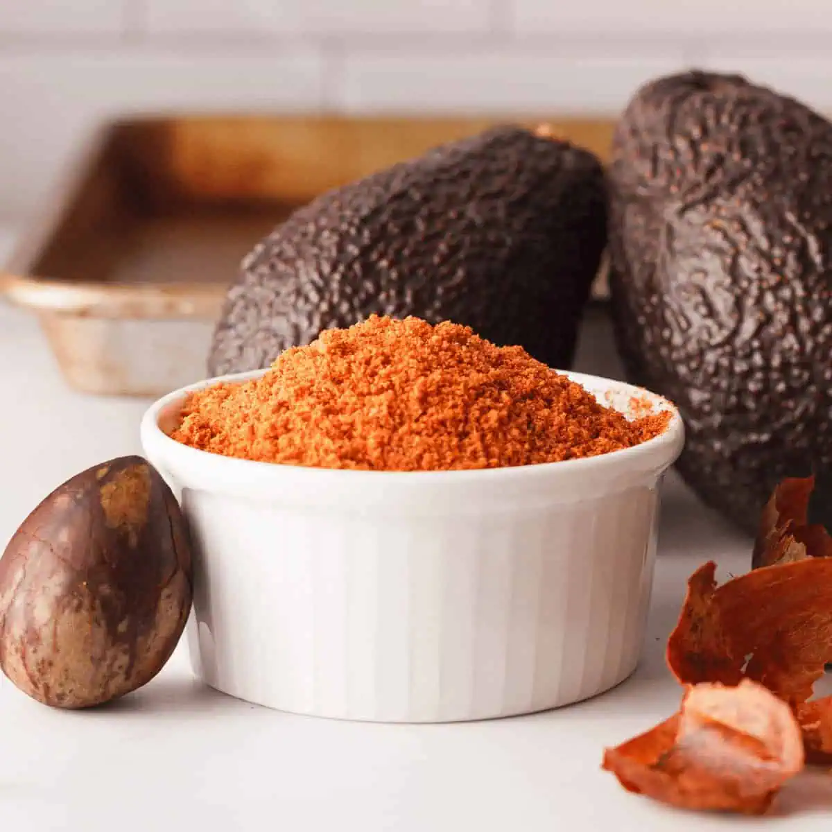 closeup of avocado seed powder in small white bowl with avocados in the background and a seed propped against the bowl