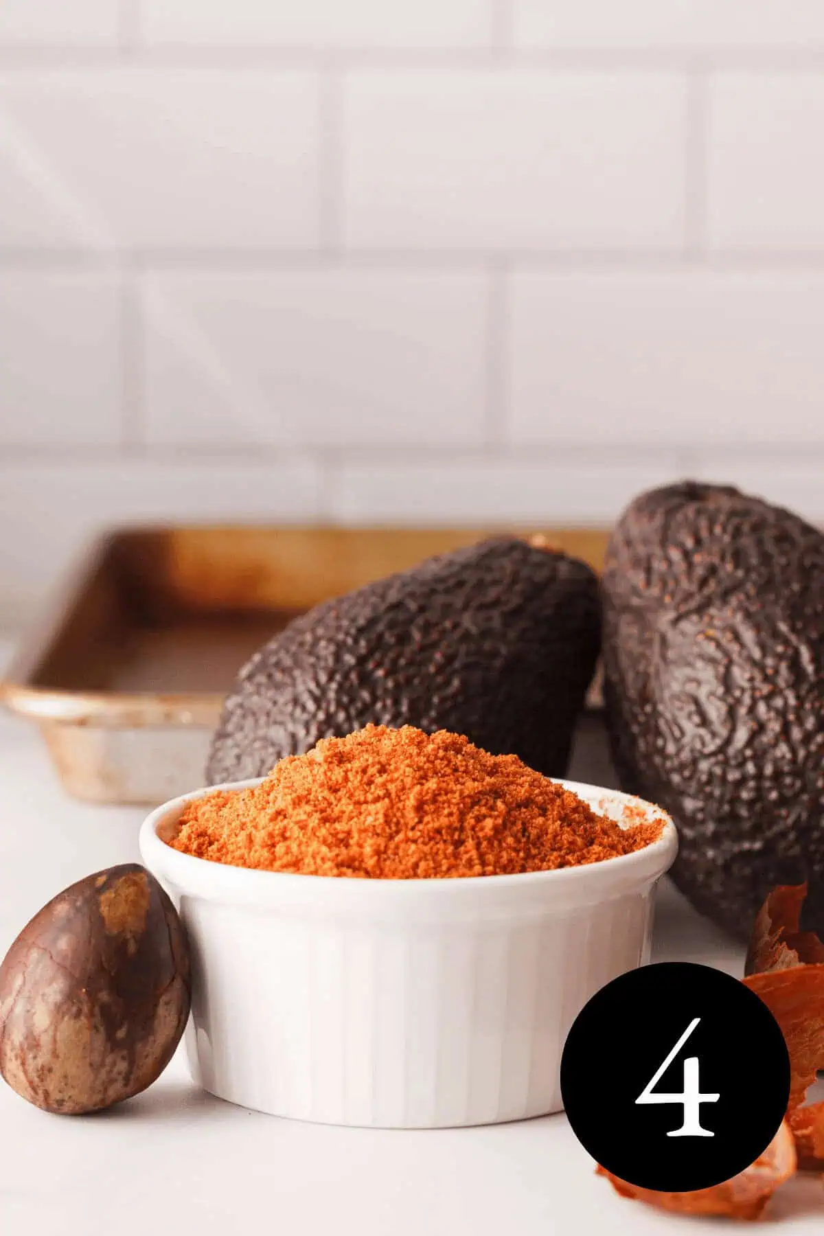 vertical closeup of ground avocado seed powder in small white dish with intact seed next to bowl and whole avocados in the background. number 4 is in the bottom right corner