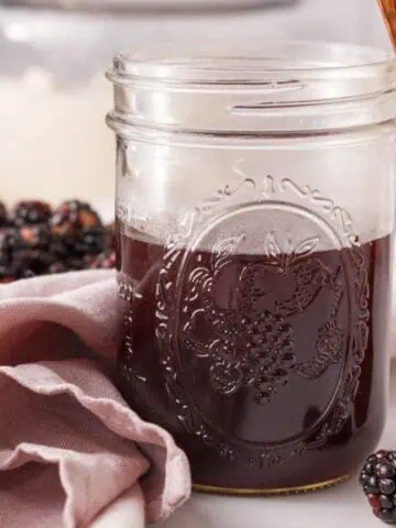 square image of mason jar half full of blackberry syrup with blackberries in foreground and background