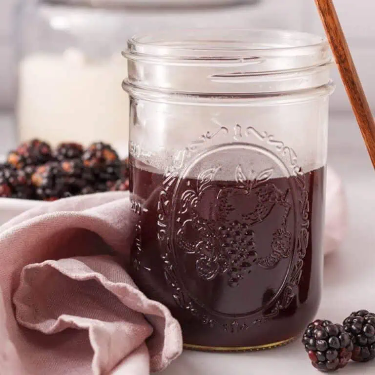 square image of mason jar half full of blackberry syrup with blackberries in foreground and background