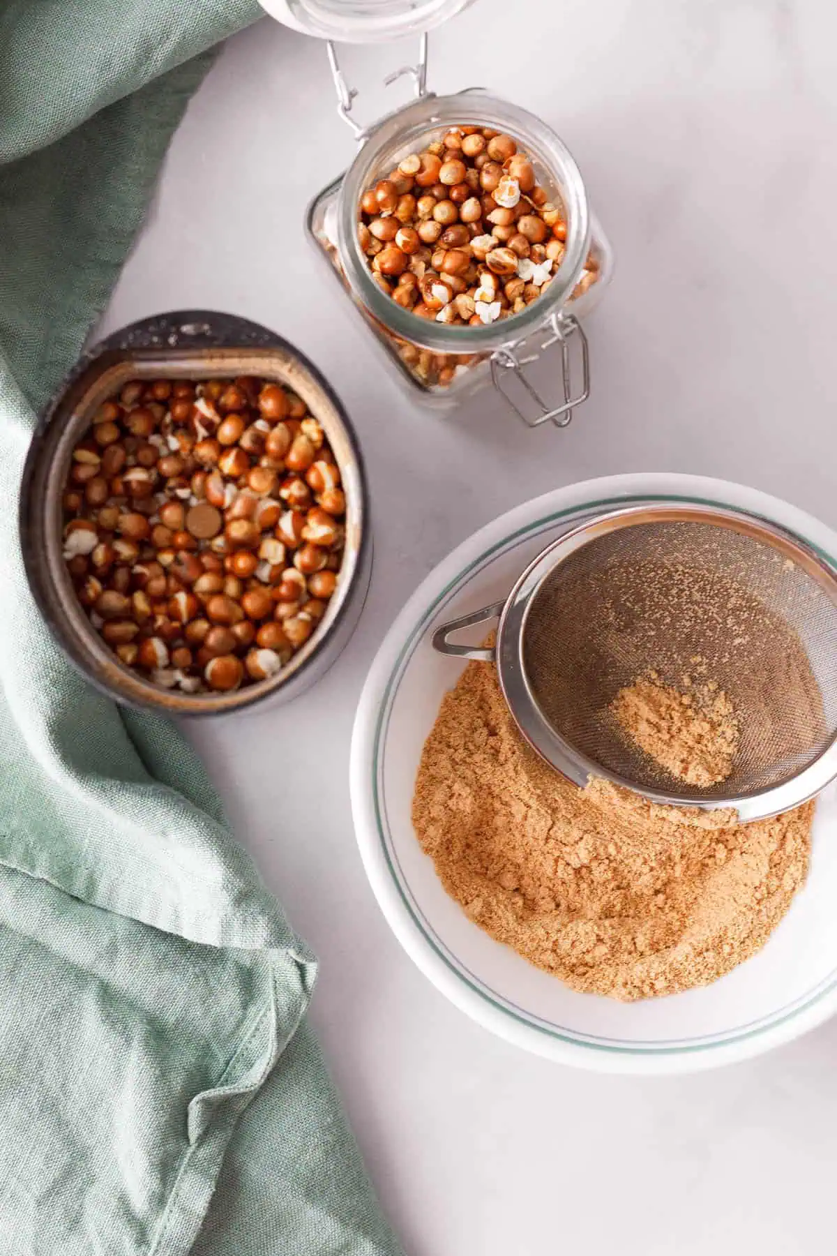 overhead image of popcorn kernels in container, spice grinder, and flour in bowl with sifter on top to show the flour-making process