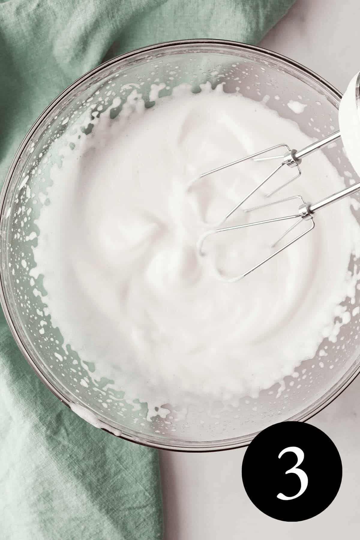overhead image of whipped aquafaba in glass bowl with beaters resting against side and number three in the bottom right corner