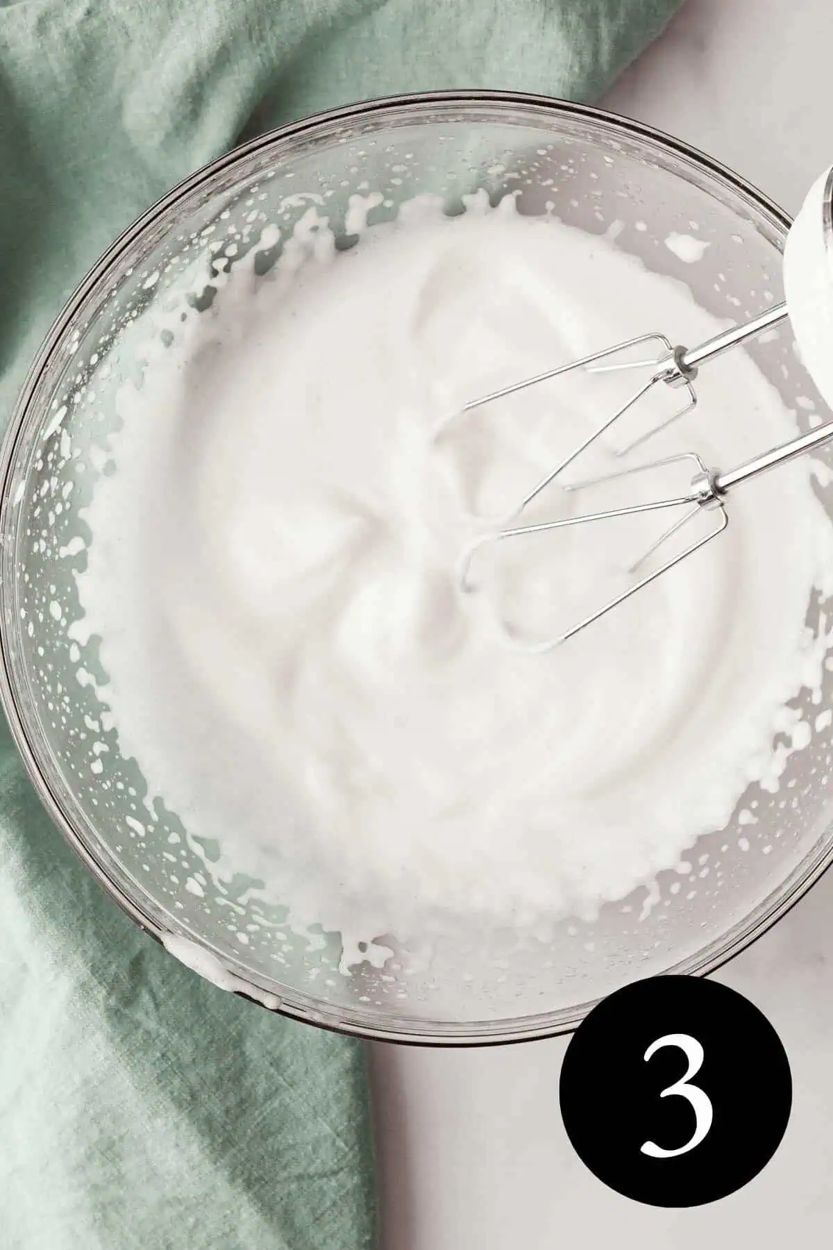 overhead image of whipped aquafaba in glass bowl with beaters resting against side and number three in the bottom right corner