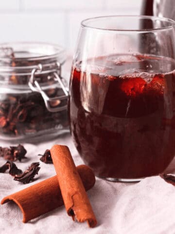 closeup of hibiscus tea with cinnamon in glass with ice with ingredients and larger carafe surrounding the glass