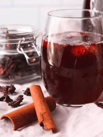 closeup of hibiscus tea with cinnamon in glass with ice with ingredients and larger carafe surrounding the glass