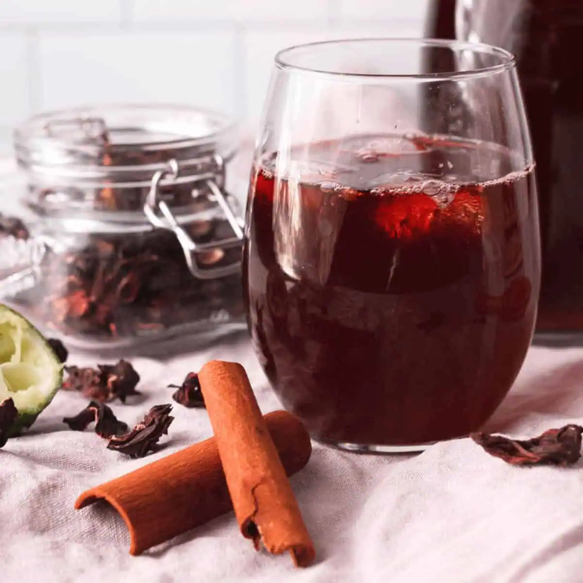 closeup of hibiscus tea with cinnamon in glass with ice with ingredients and larger carafe surrounding the glass