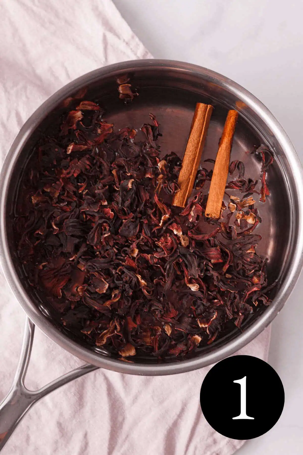 overhead image of hibiscus and cinnamon sticks in saucepan with water and number one in bottom right corner