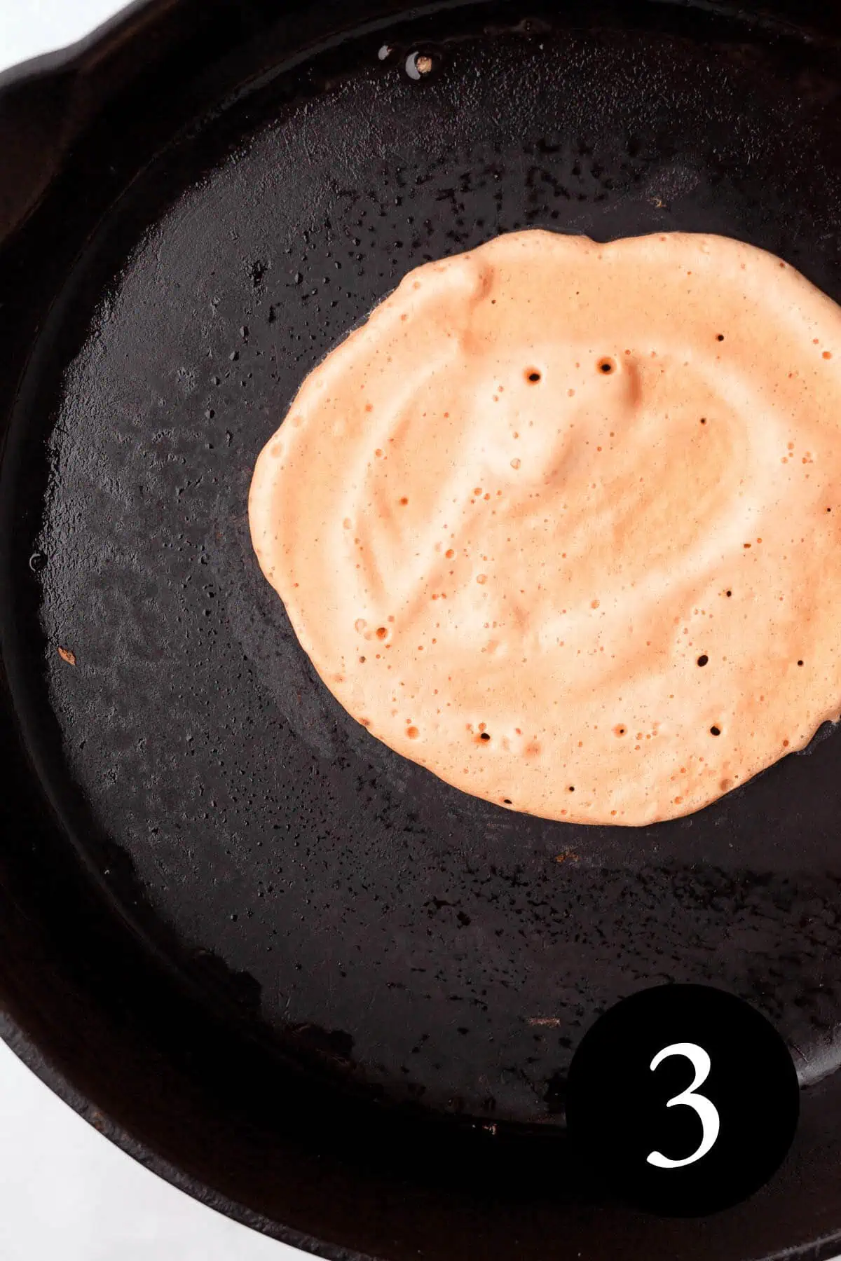 overhead image of a red lentil tortilla cooking in cast iron skillet with number three in bottom right corner