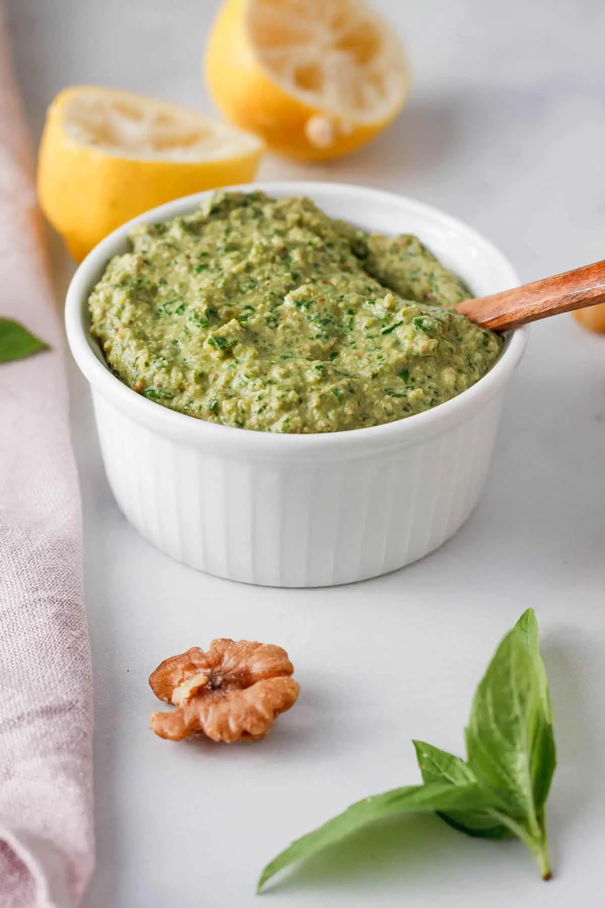 angled image of vegan basil pesto in white bowl with basil, walnuts, and used lemon halves on counter