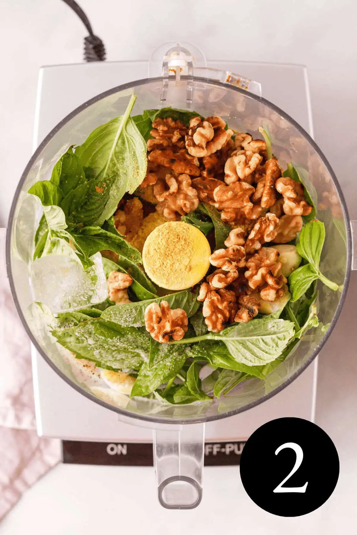 overhead image of vegan basil pesto ingredients in bowl of food processor before blending with number two in the bottom right corner
