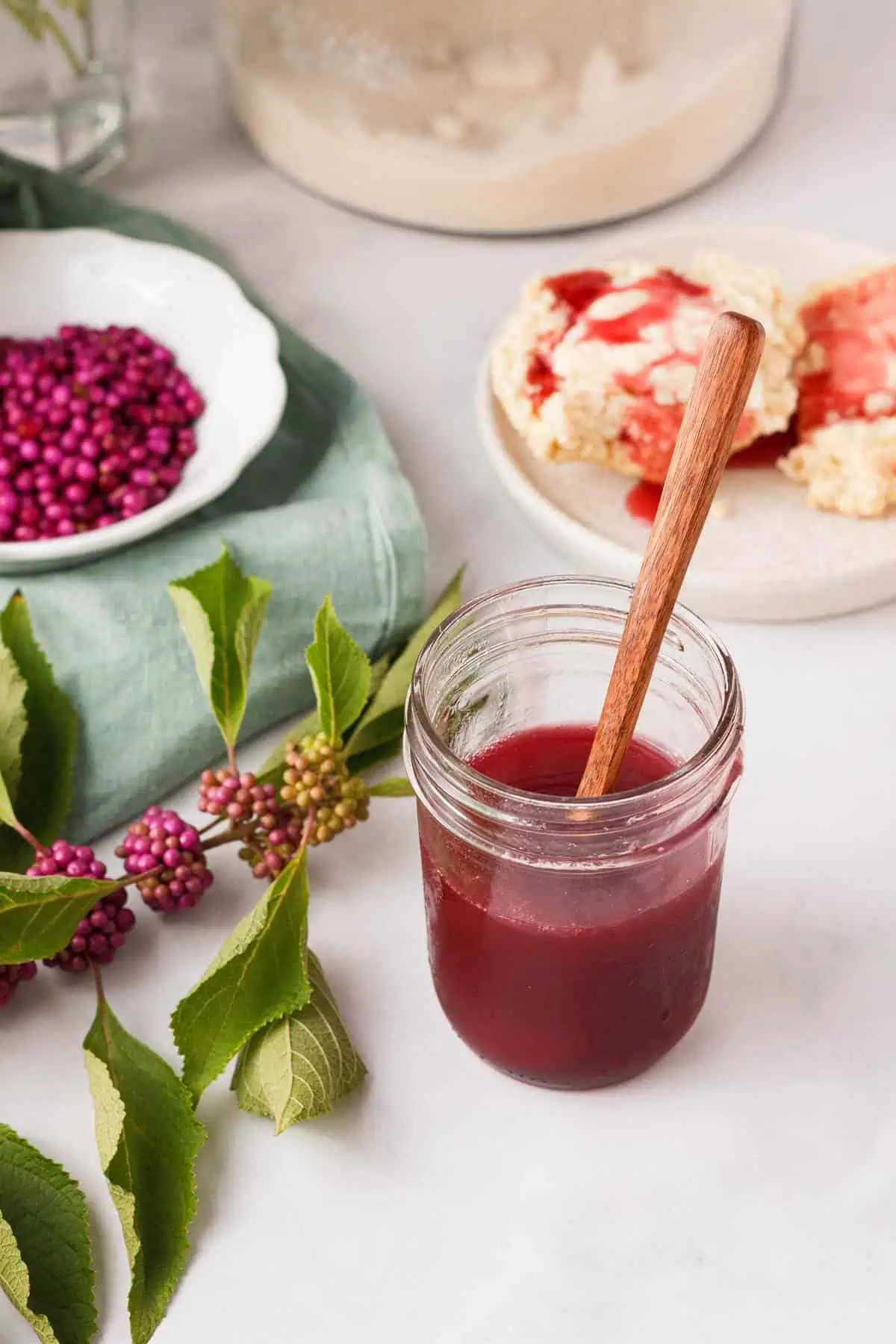 angled image of jarred beautyberry jelly next to branch, loose berries, and biscuits in background
