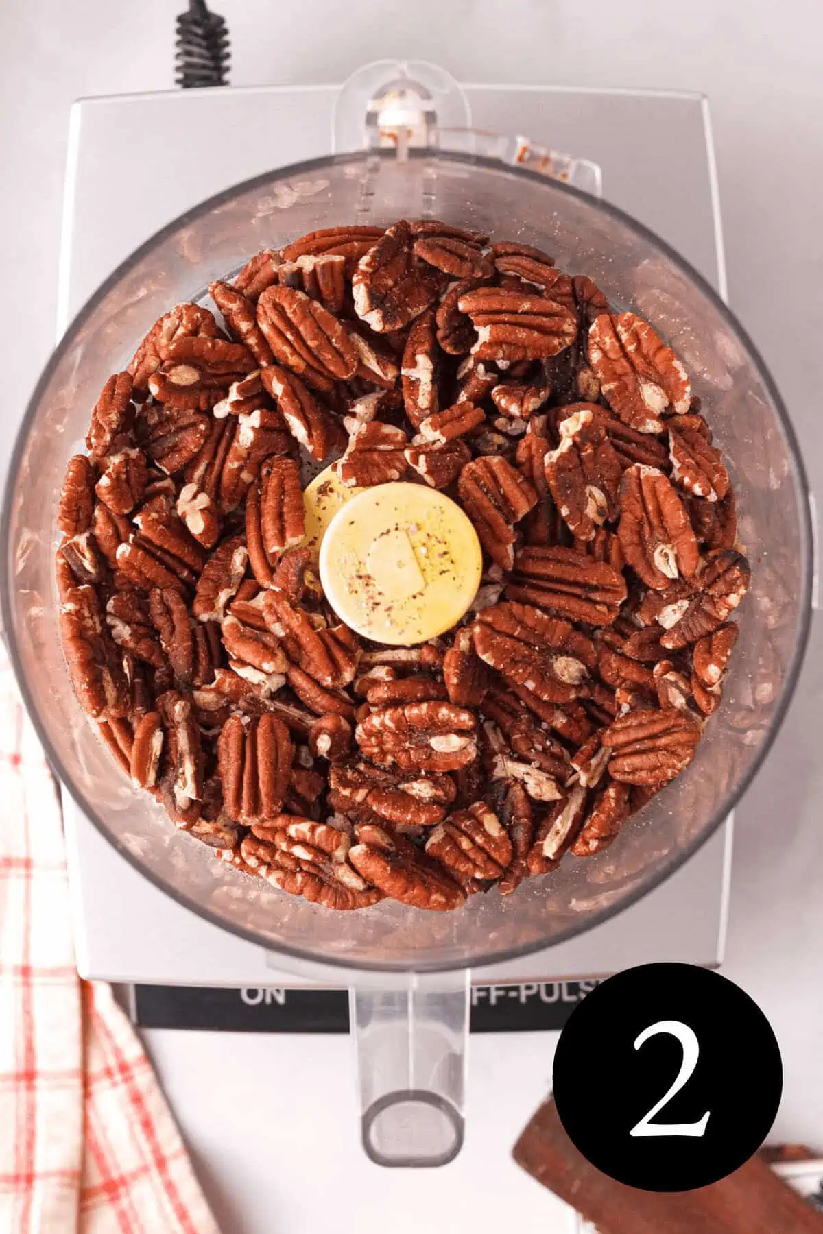 overhead image of roasted pecans in bowl of food processor and number two in the bottom right corner