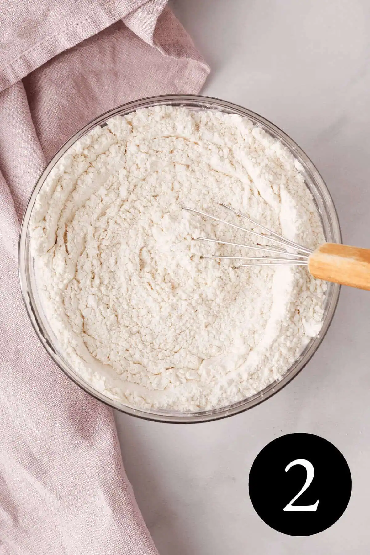 overhead image of whisked flour and baking powder in bowl with whisk resting on the edge and number two in the bottom right corner